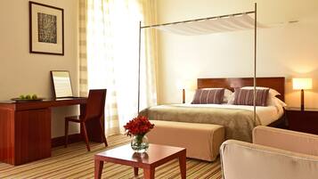 A hotel room with a bed, a desk, a chair, a small table, and a vase of flowers.