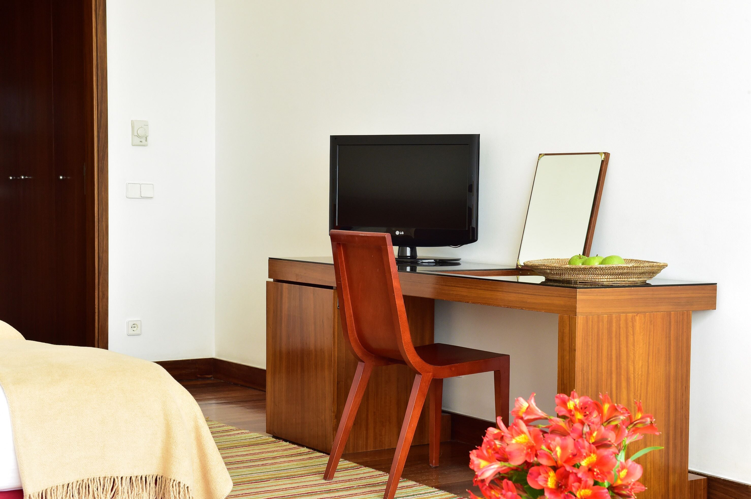 Ein Hotelzimmer mit Schreibtisch aus Holz, einem Fernseher, einem Spiegel und einem kleinen Tisch mit Obstkorb.