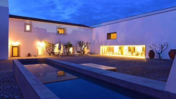 Un patio con piscina, mobiliario exterior y un edificio con ventanas iluminadas.