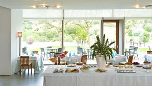 Comedor con mesas dispuestas para un bufé, amplias ventanas con vista a follaje y luces en el techo.