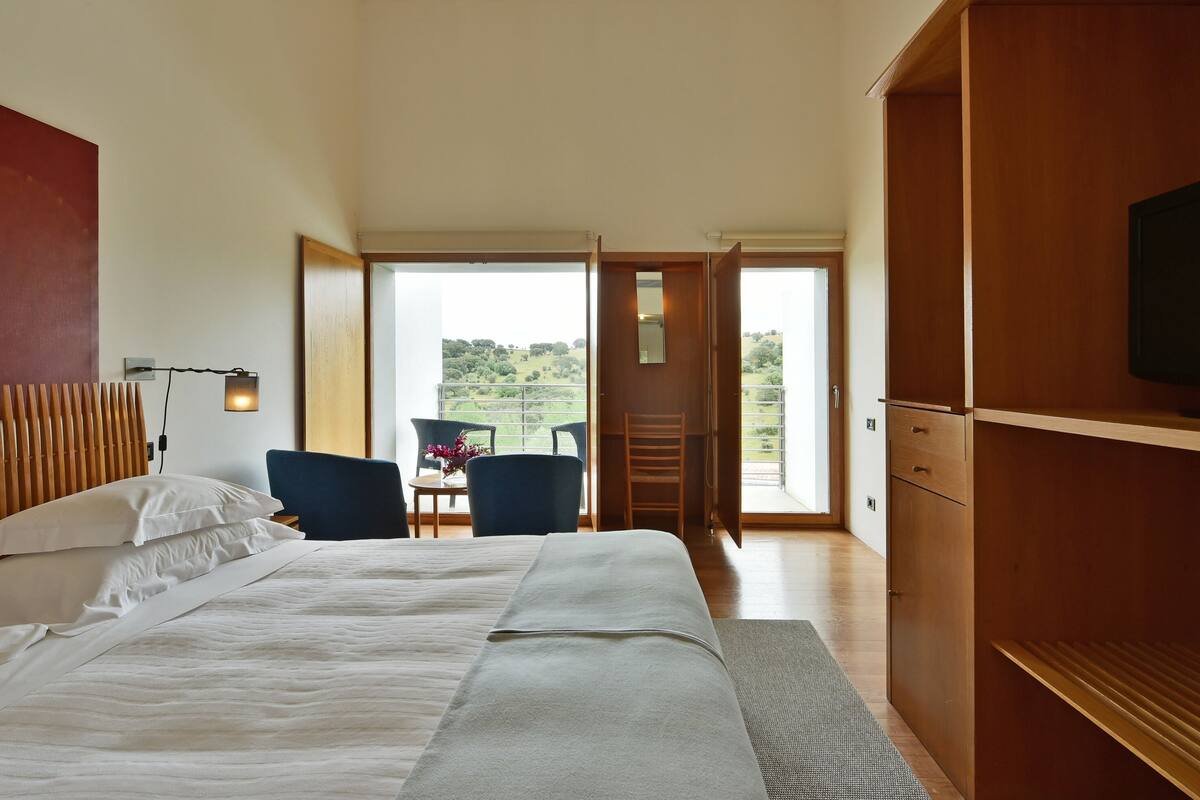 A hotel room with a large bed, a desk with a chair, a TV mounted on the wall, and a balcony with a view of the countryside.