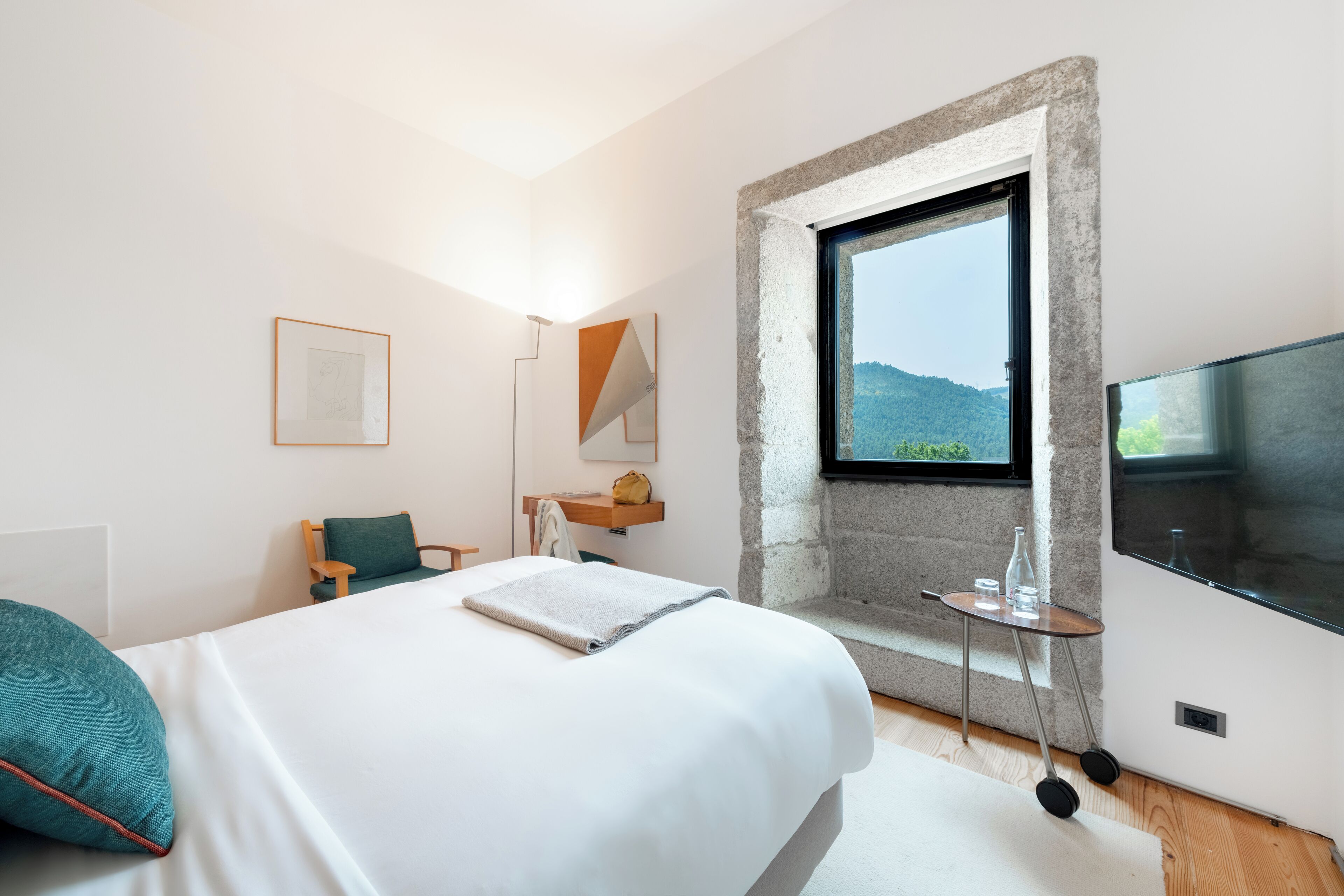 A modern hotel room with a large bed, a small table, a TV, and a window with a mountain view.