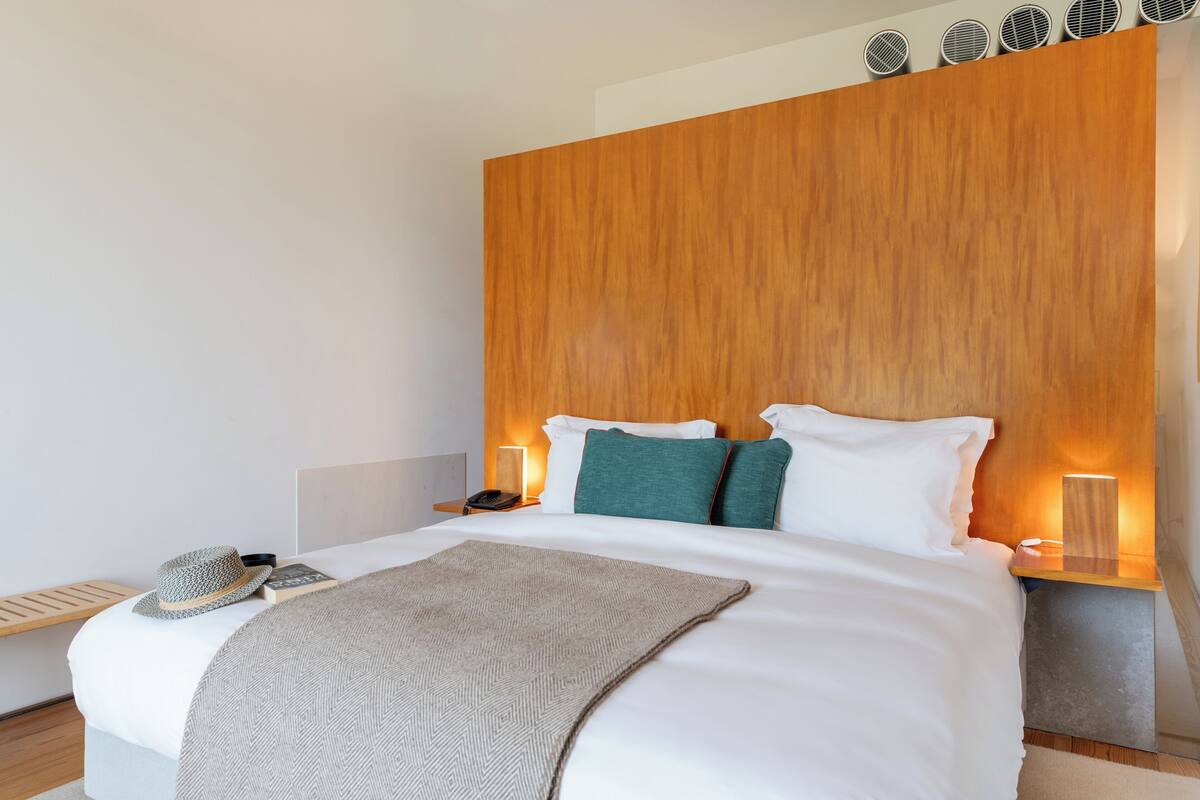 Una habitación de hotel moderna con cabecera de madera, ropa de cama blanca, una lámpara de noche, un sombrero y un libro sobre la mesita de noche.