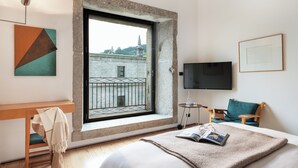 Un cuarto de hotel con un ventanal grande, una cama con manta, una silla, una mesita con vasos y un televisor en la pared.