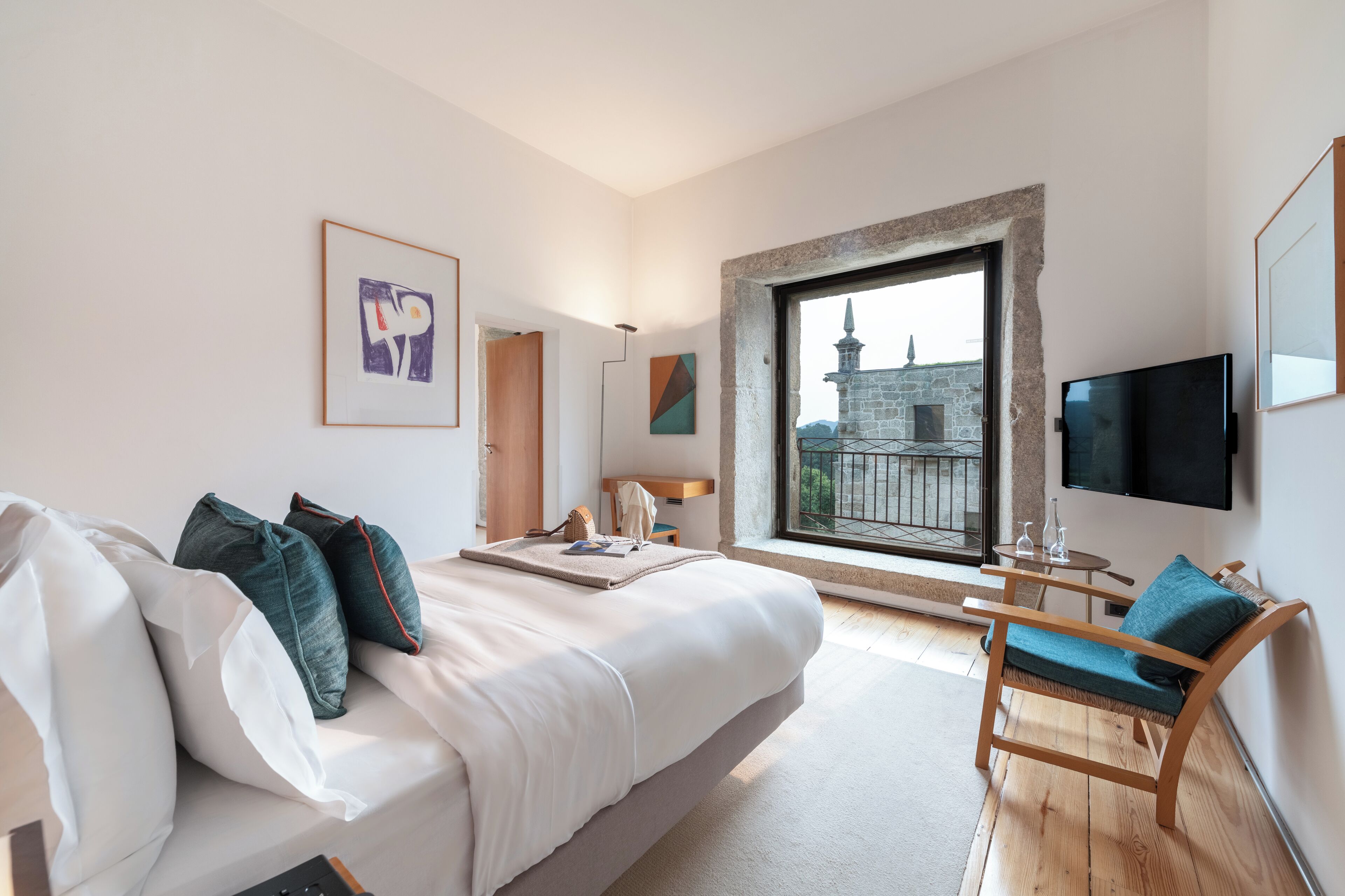 Un dormitorio moderno con una cama grande, una silla de madera, un televisor y una ventana con vistas a un edificio.
