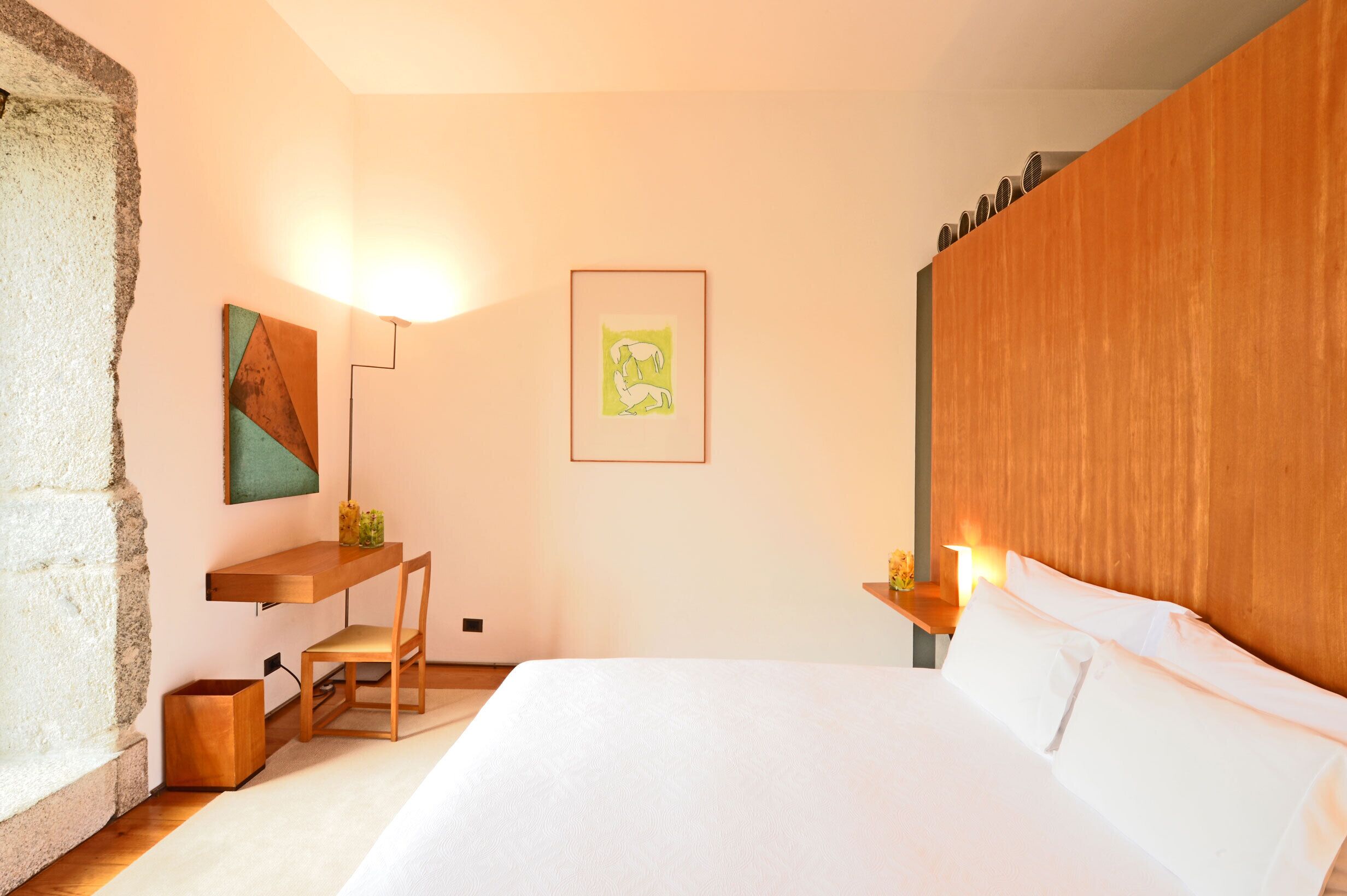 A bedroom with a bed, a desk, a chair, a wooden headboard, and artwork on the walls.