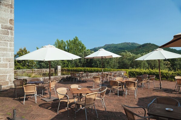 An outdoor patio with wicker furniture and white umbrellas, overlooking a landscaped garden and hills. - Pousada Mosteiro Amares (Amares)