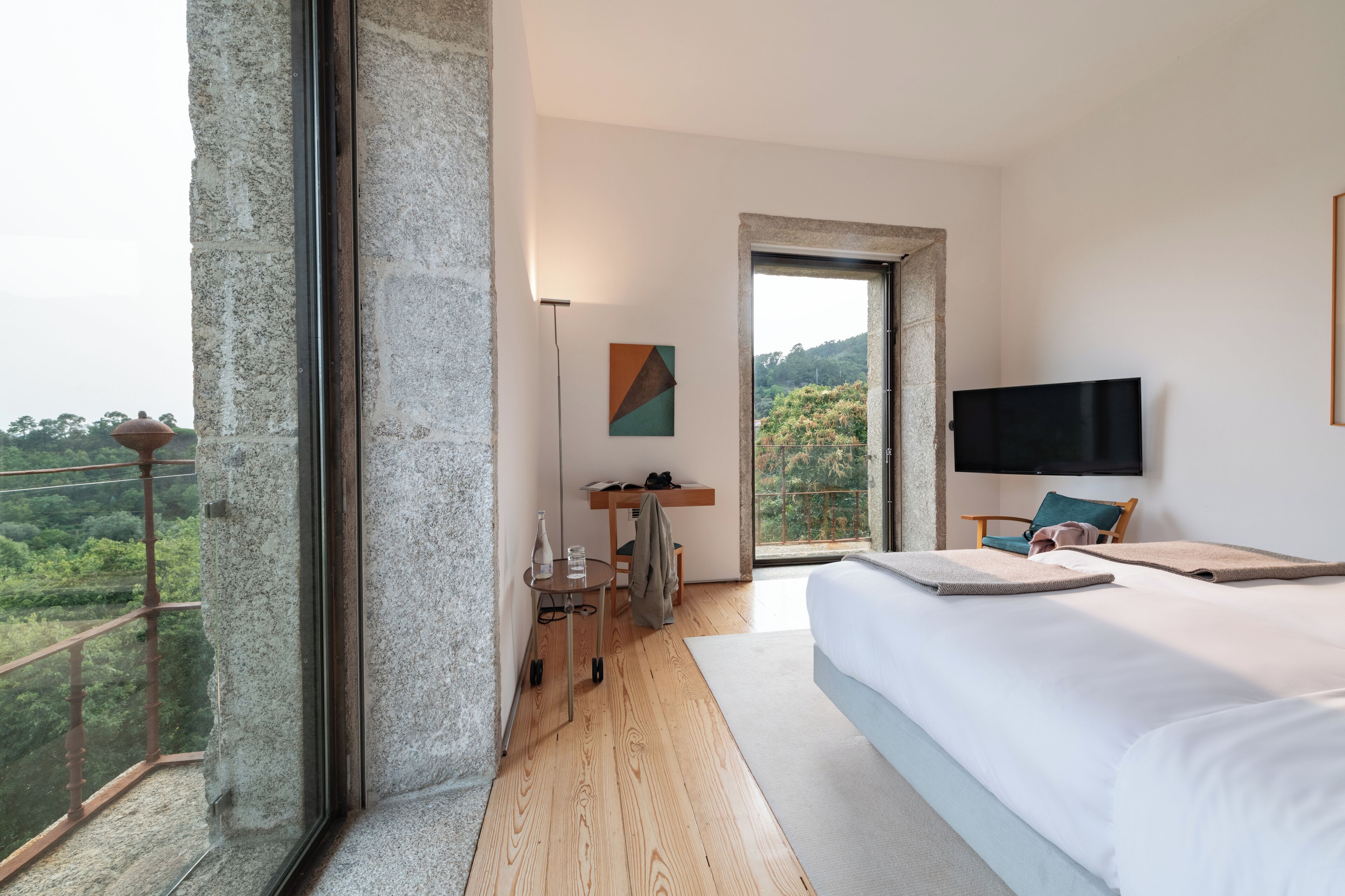 Une chambre d’hôtel moderne dotée d’un grand lit, d’une télévision, d’un bureau avec une chaise et d’un balcon offrant une vue sur la forêt.