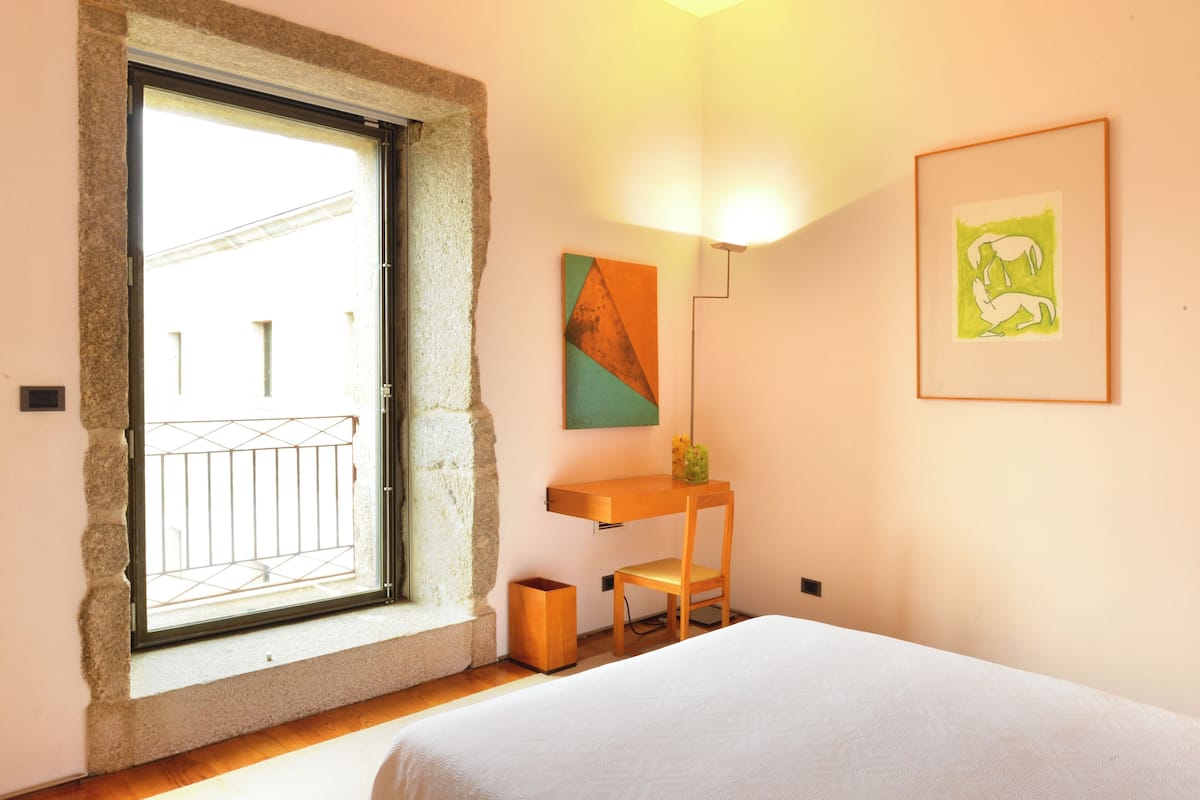 Un dormitorio con cama, escritorio, silla, ventana y obras de arte en las paredes.