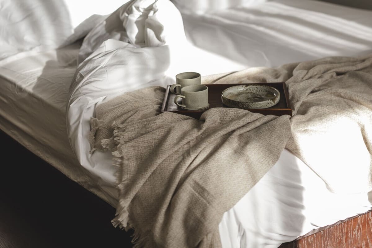 A bed with white linens, a wooden tray with a cup and saucer, and a decorative plate.