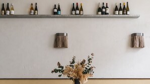Un comedor con una mesa redonda, cuatro sillas de madera, un florero con flores secas y una estantería para vinos.