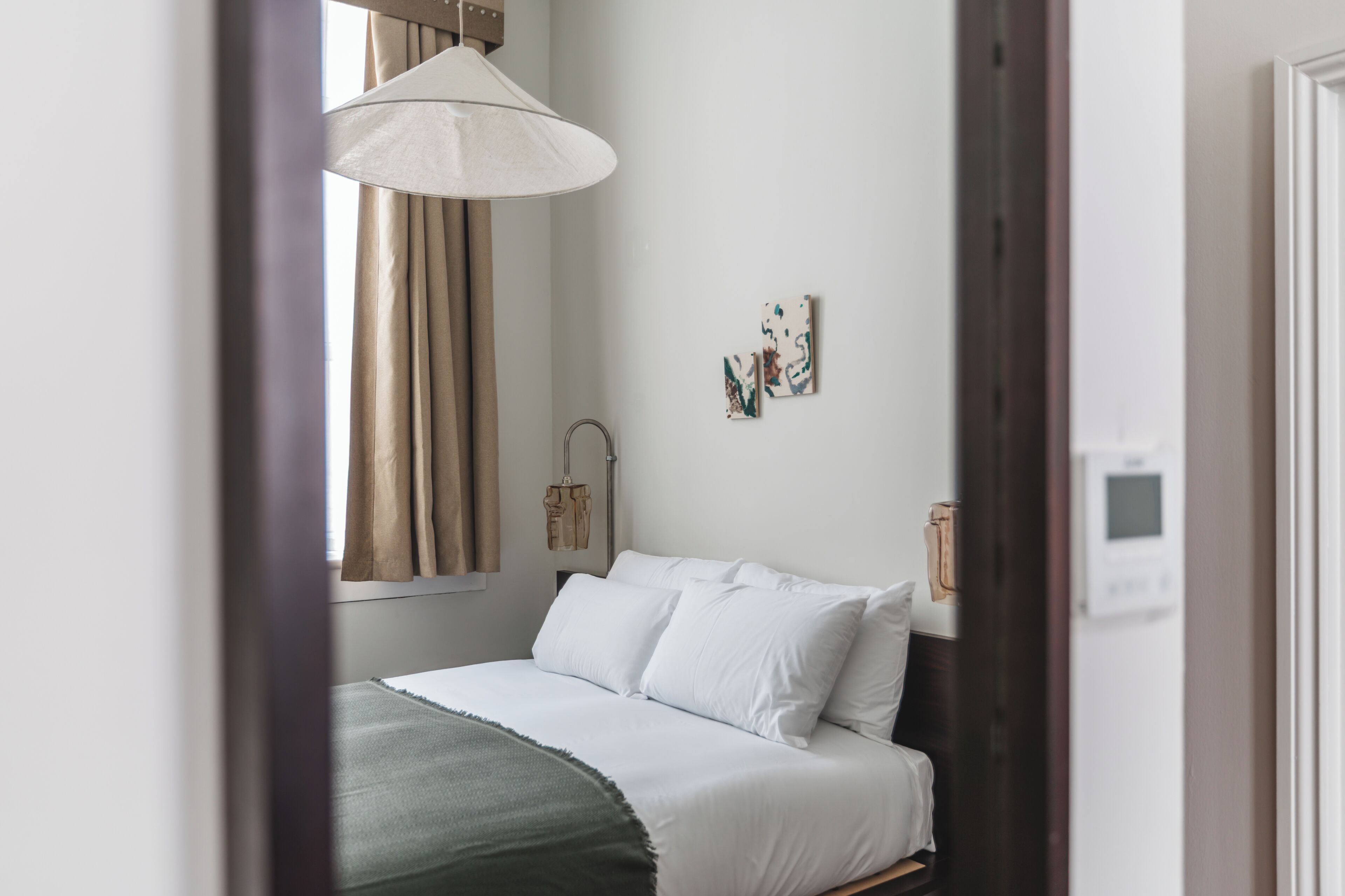 A bedroom with a bed, a window with curtains, a wall-mounted light fixture, and a thermostat.