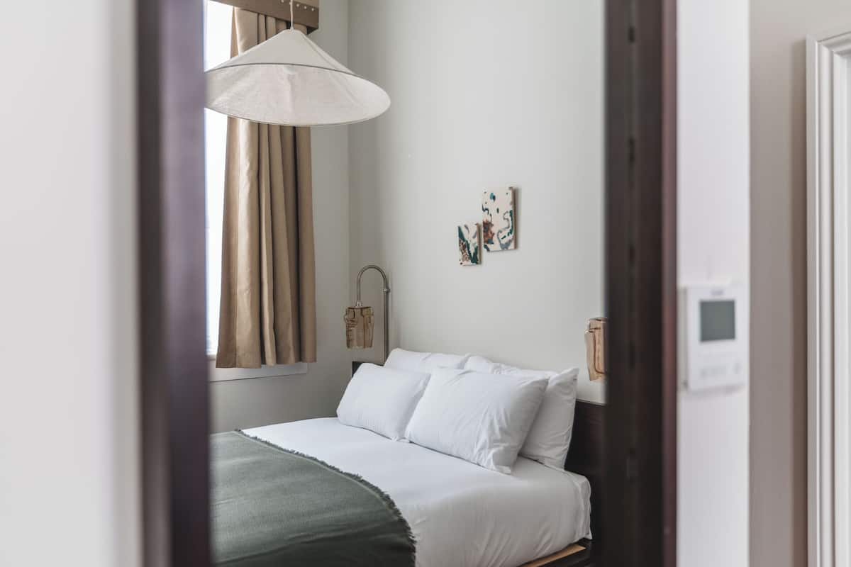 A bedroom with a bed, a window with curtains, a wall-mounted light fixture, and a thermostat.