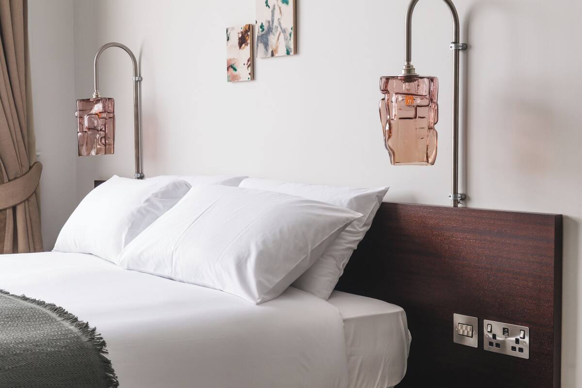 A neatly made bed with white linens, two wall-mounted reading lamps, and a small wall outlet.
