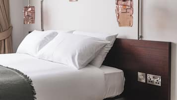 Cama bem arrumada com lençóis brancos, duas luminárias de parede para leitura e uma pequena tomada na parede.