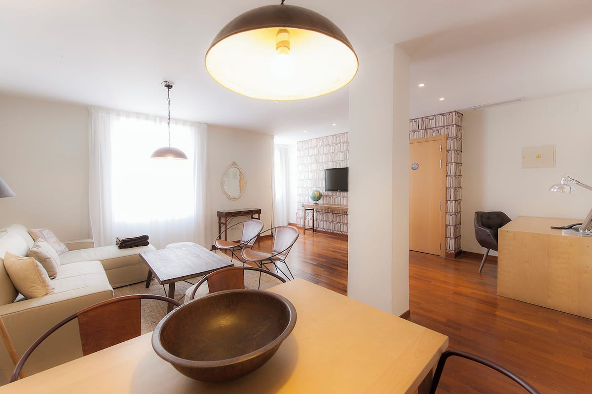 Una sala moderna con sofá, mesa de comedor y sillas. En la mesa hay un cuenco de madera grande, una lámpara colgante y una estantería llena de libros.