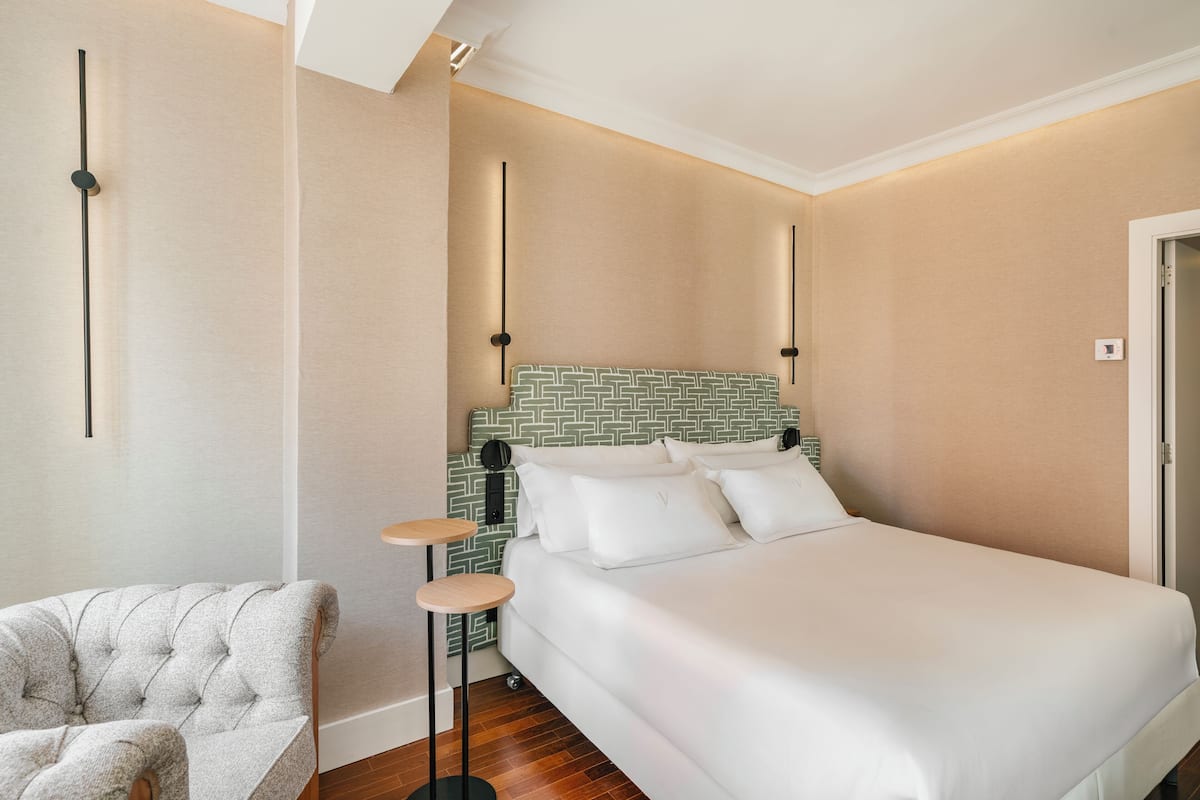 Un dormitorio ordenado con una cama grande, un sillón capitoné y una mesita de noche pequeña.
