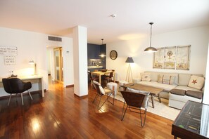 A modern living room with a wooden floor, a dining area with a table and chairs, a sofa, and a kitchen in the background. - Hotel Vincci SoMa (Madrid)