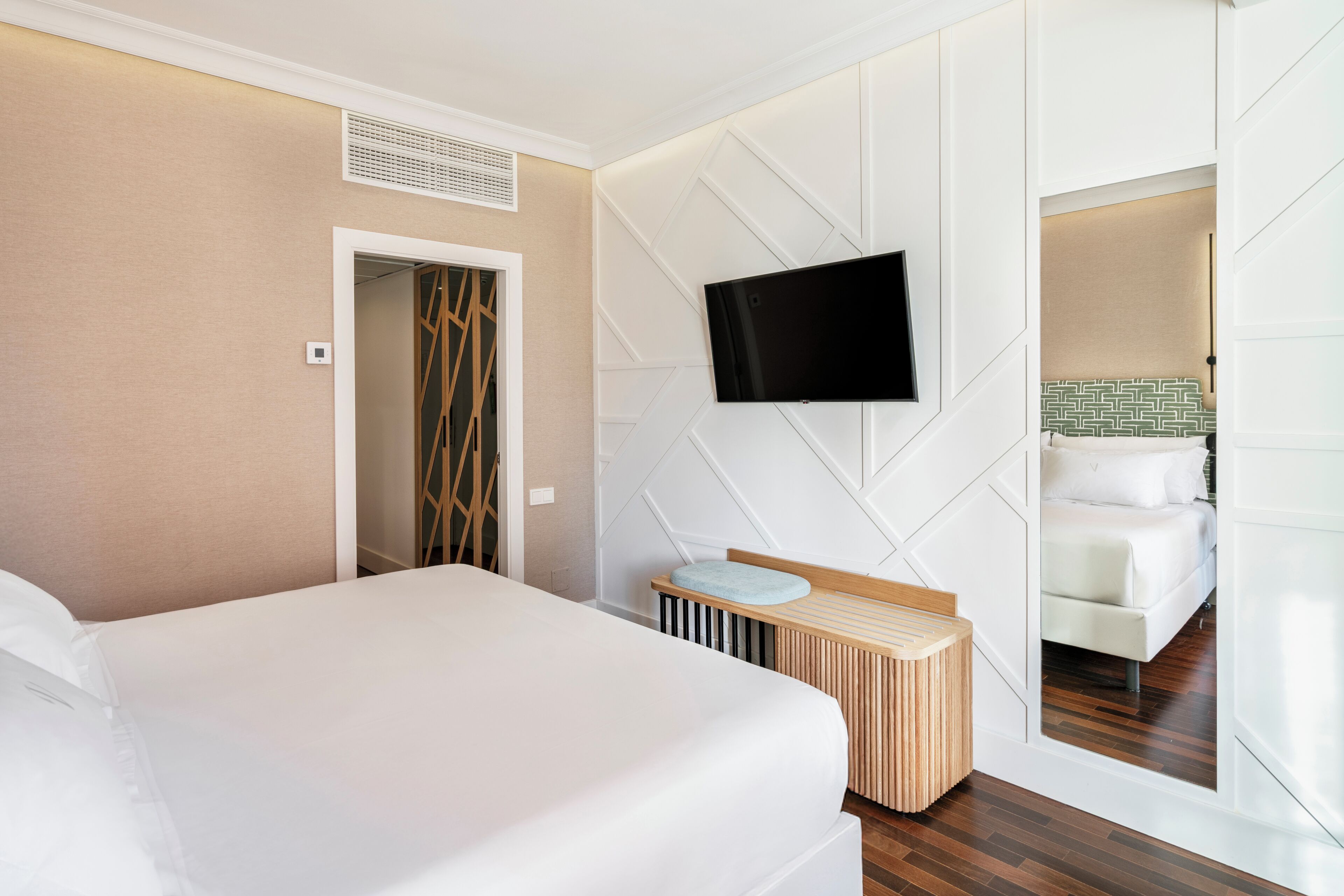 Quarto de hotel com uma cama, televisão fixada na parede, uma pequena mesa de cabeceira de madeira e um espelho refletindo uma cama e um banheiro.