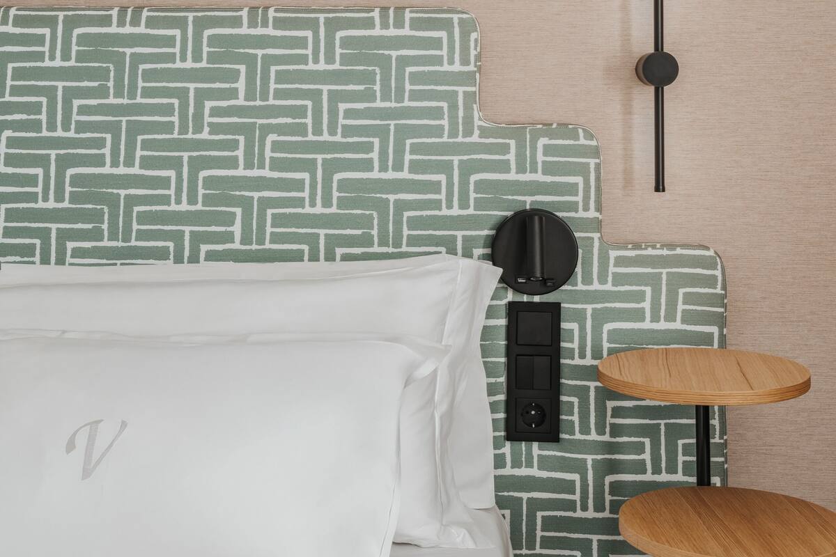 Una cama con cabecera estampada, una mesita de noche y un aplique de pared.