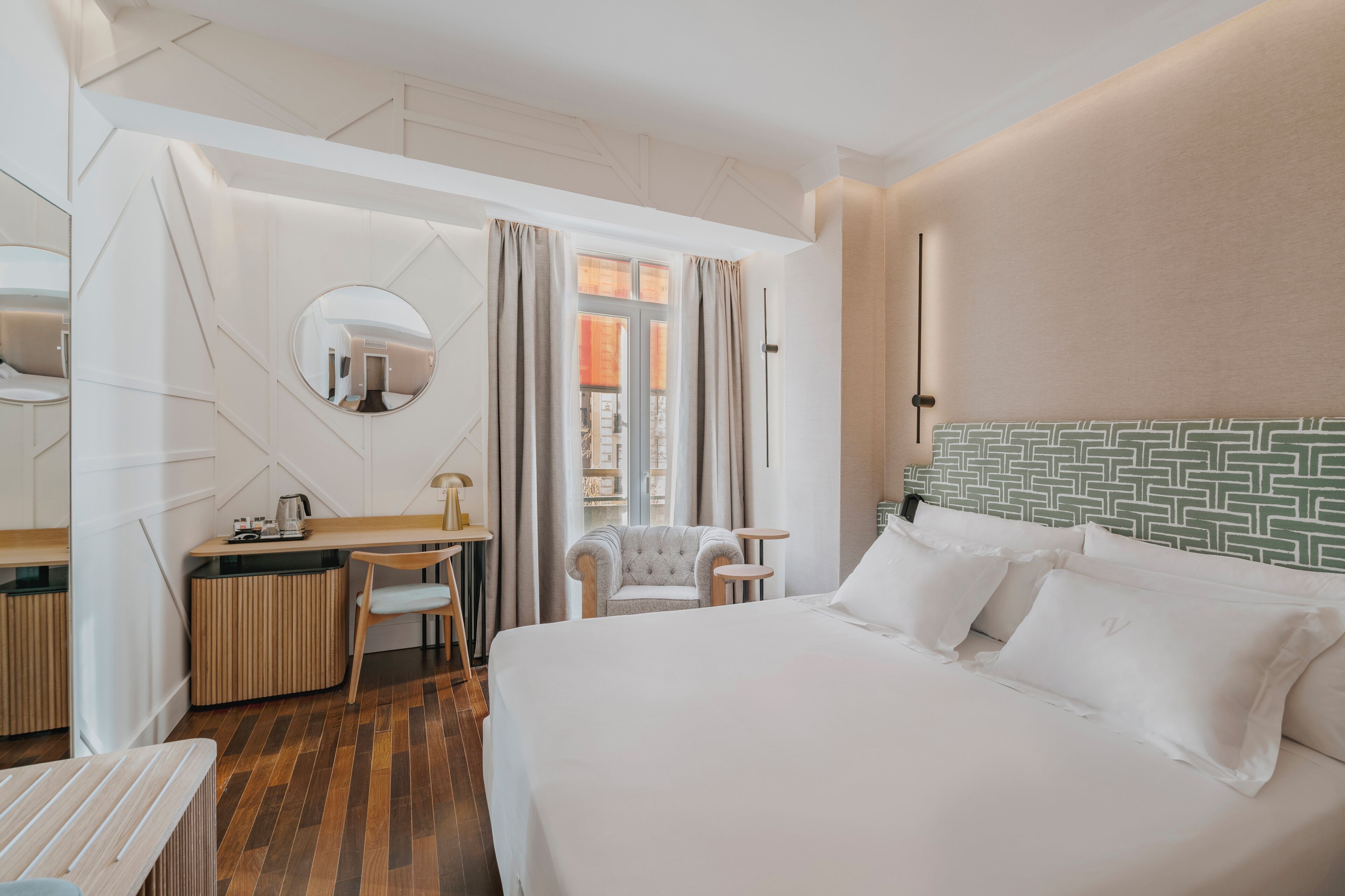 A hotel room with a bed, a desk with a chair, a round mirror, and a view of the city through the window.