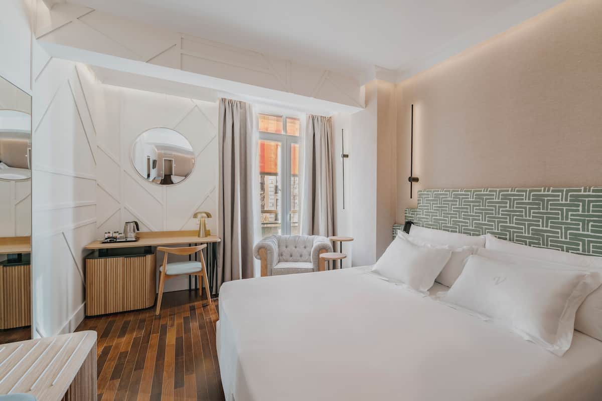 Habitación de hotel con una cama, un escritorio con silla, un espejo redondo y vistas a la ciudad desde la ventana.