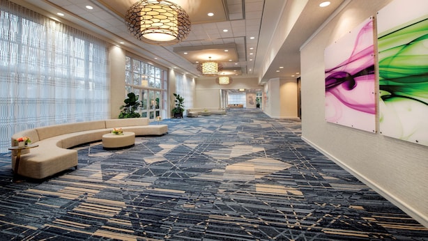 Un hall d’hôtel spacieux, avec un canapé incurvé, une moquette à motifs et une grande fenêtre ornée de rideaux légers.
