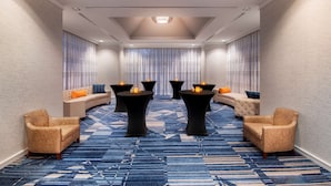 Un hall d’hôtel doté d’un tapis à motifs bleus, de tables rondes noires ornées de bougies, de canapés beiges et d’un espace salon aux lignes courbes.