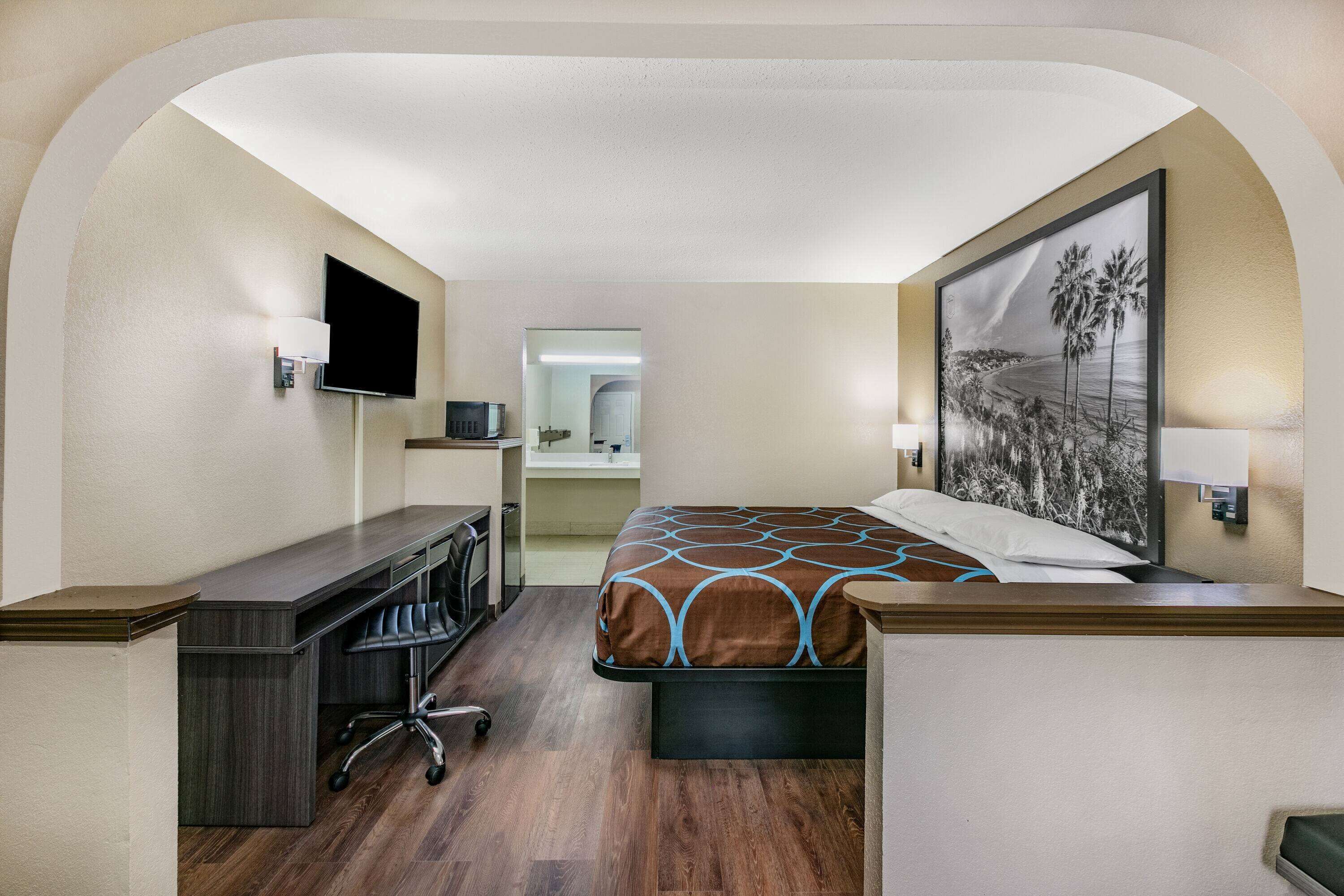 A hotel room with a bed, desk, chair, TV, and artwork on the wall.