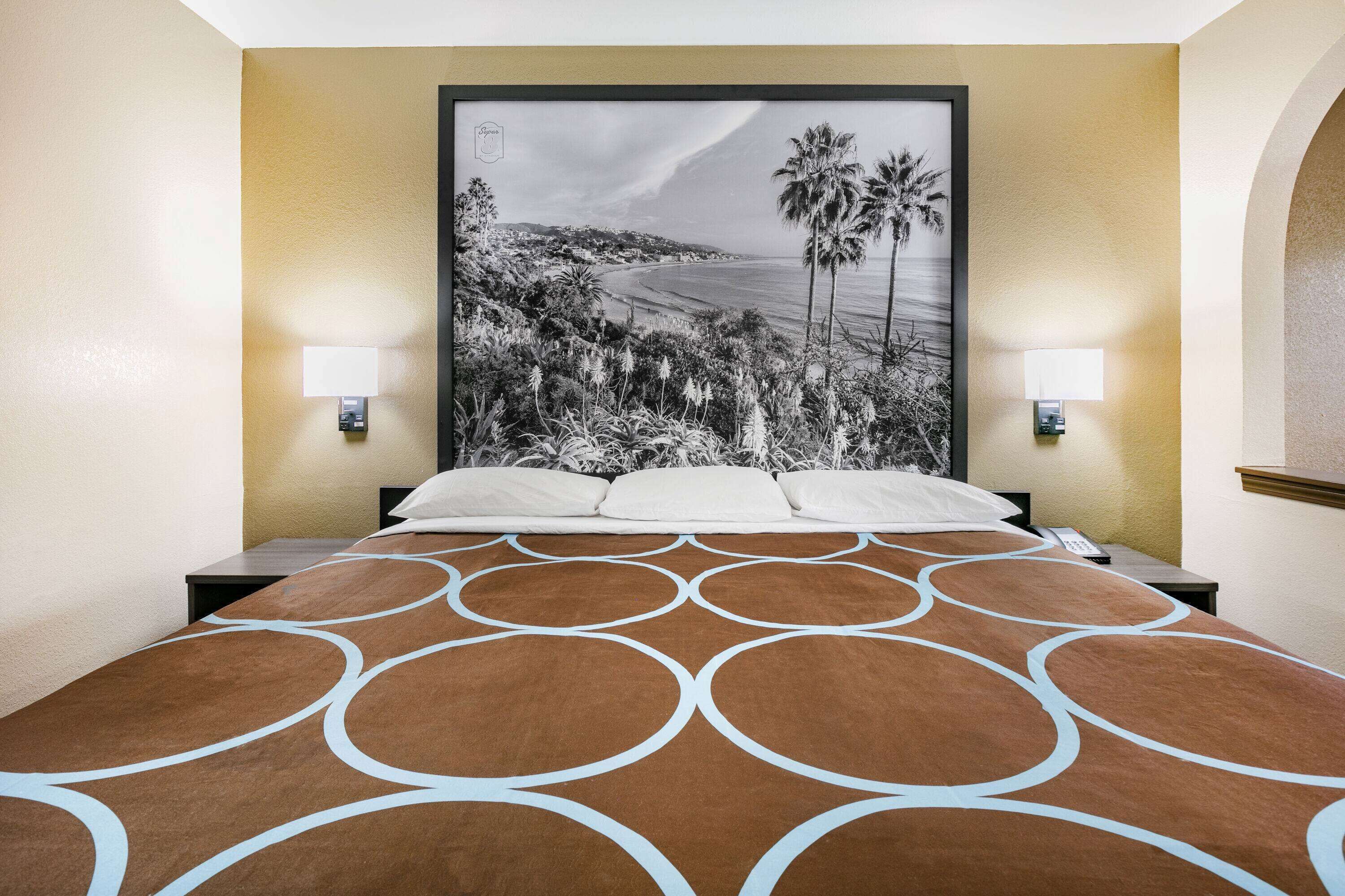 A hotel room with a large bed, a headboard with a black and white photograph of a coastal landscape, two bedside lamps, and a brown patterned bedspread.