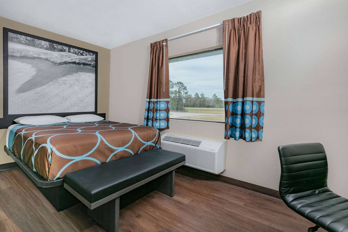 A hotel room with a bed, a bench, a chair, a large window with curtains, an air conditioning unit, and a wall-mounted artwork.
