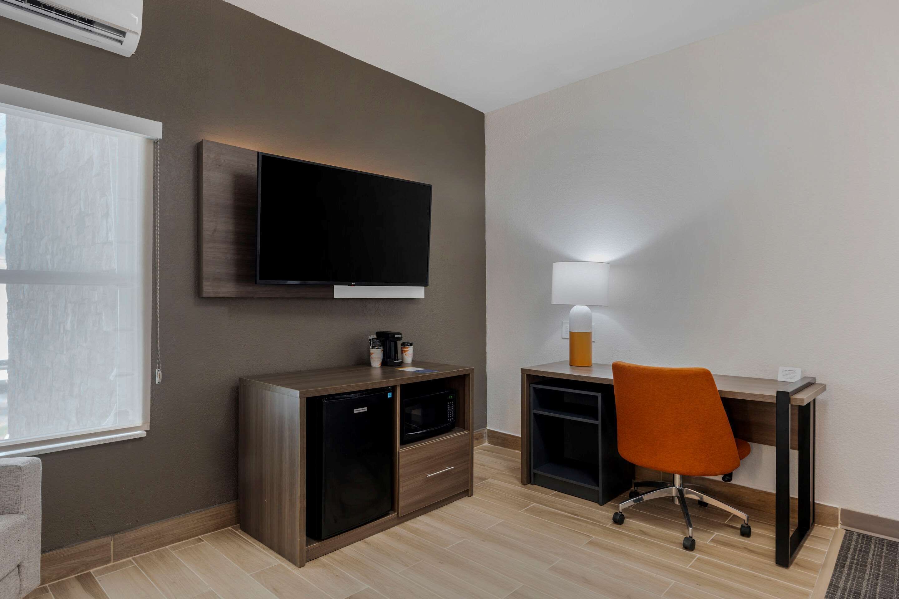 A modern hotel room with a flat-screen TV mounted on the wall, a compact kitchenette with a microwave, and a desk with an orange office chair.