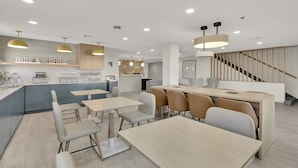 A modern kitchen with a central island, a dining area with light wood tables and chairs, and a staircase with wooden railings.