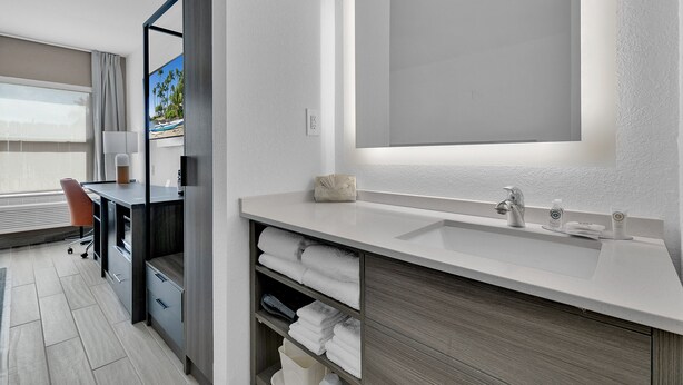 A modern bathroom with a large mirror, a sink, and a desk with a chair. There are towels and toiletries on the counter.