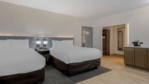 A hotel room with two beds, a desk, a chair, a small kitchenette, and a door leading to a bathroom.