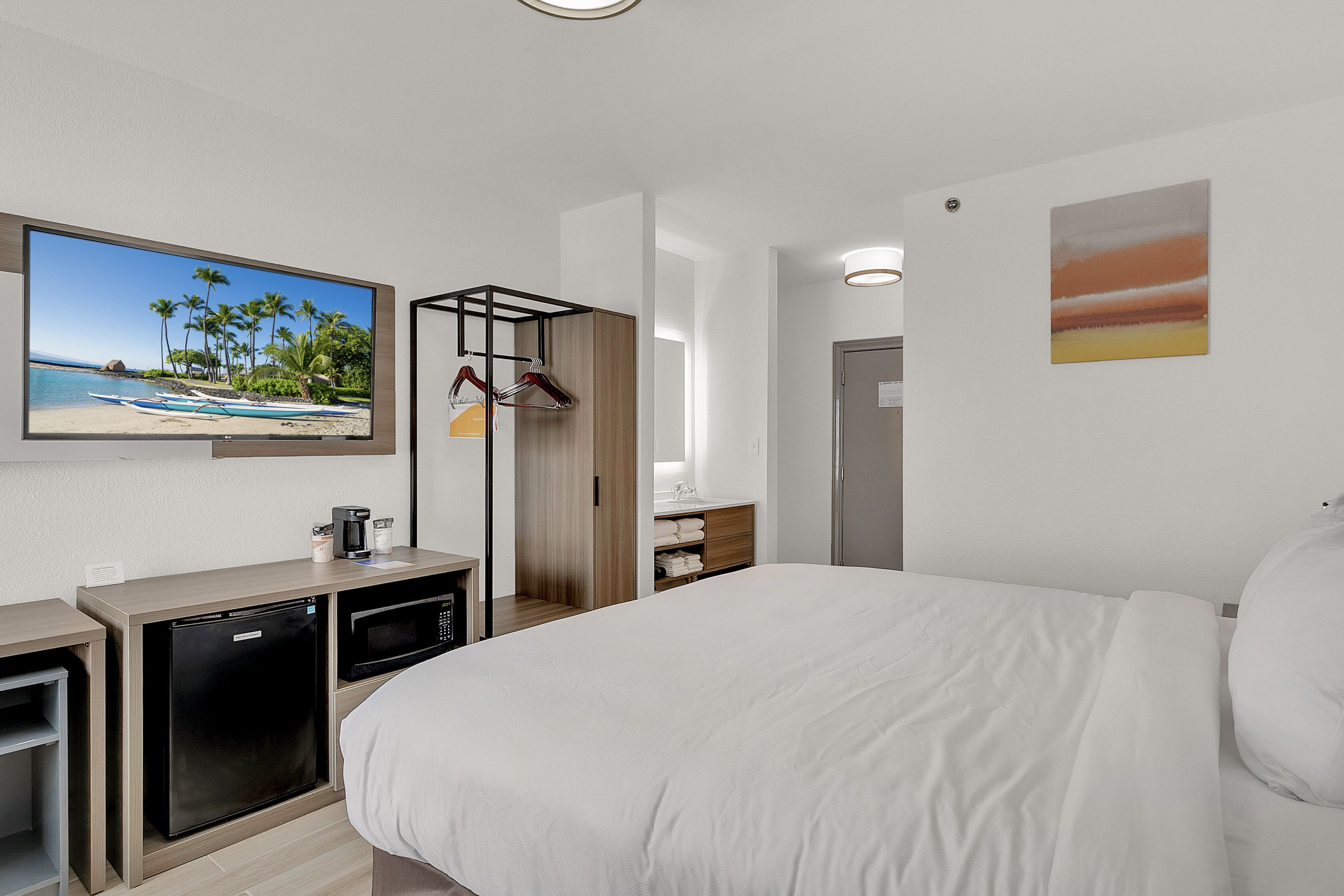 Habitación de hotel con cama, escritorio con refrigerador y microondas, televisor mostrando una escena de playa y armario con ropa colgada.