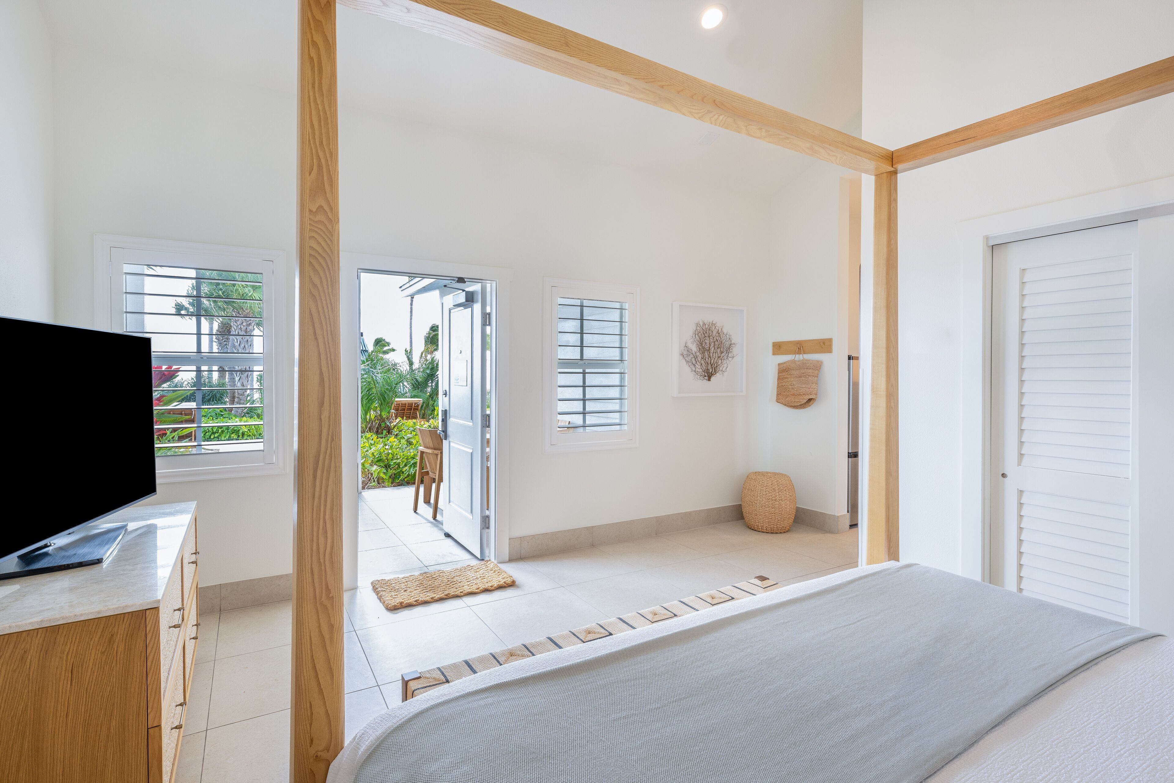 A modern bedroom with a bed, a TV on a wooden dresser, and a view of a garden through an open door.