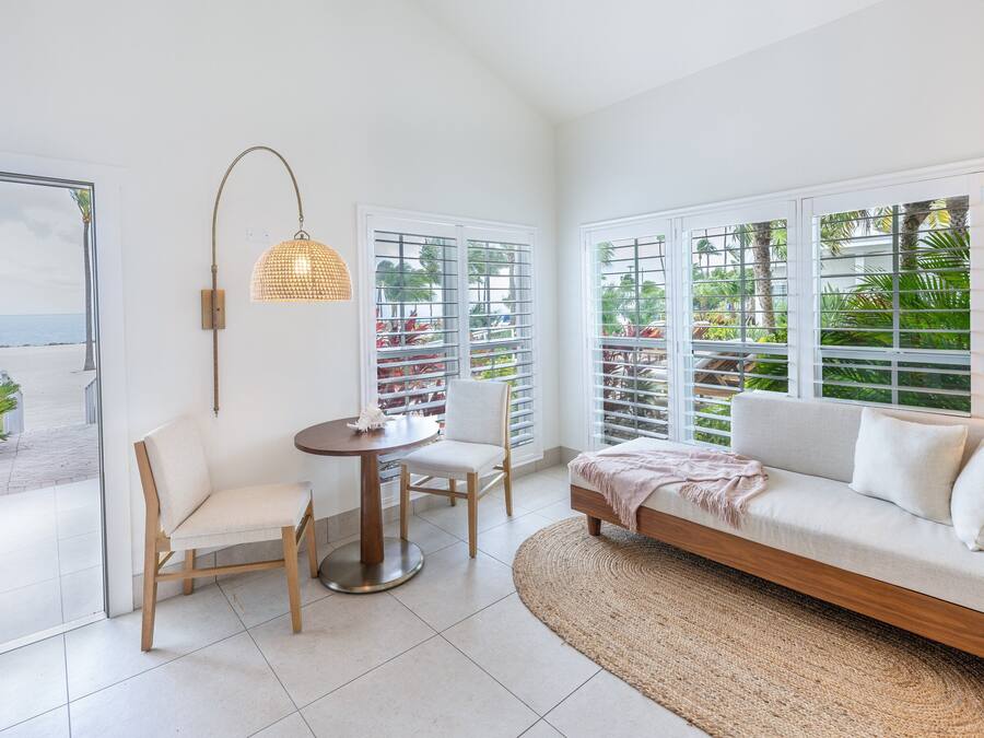 A modern living room with a white sofa, a round table, and a chair. There are large windows with white blinds, a woven pendant light, and a view of the beach.