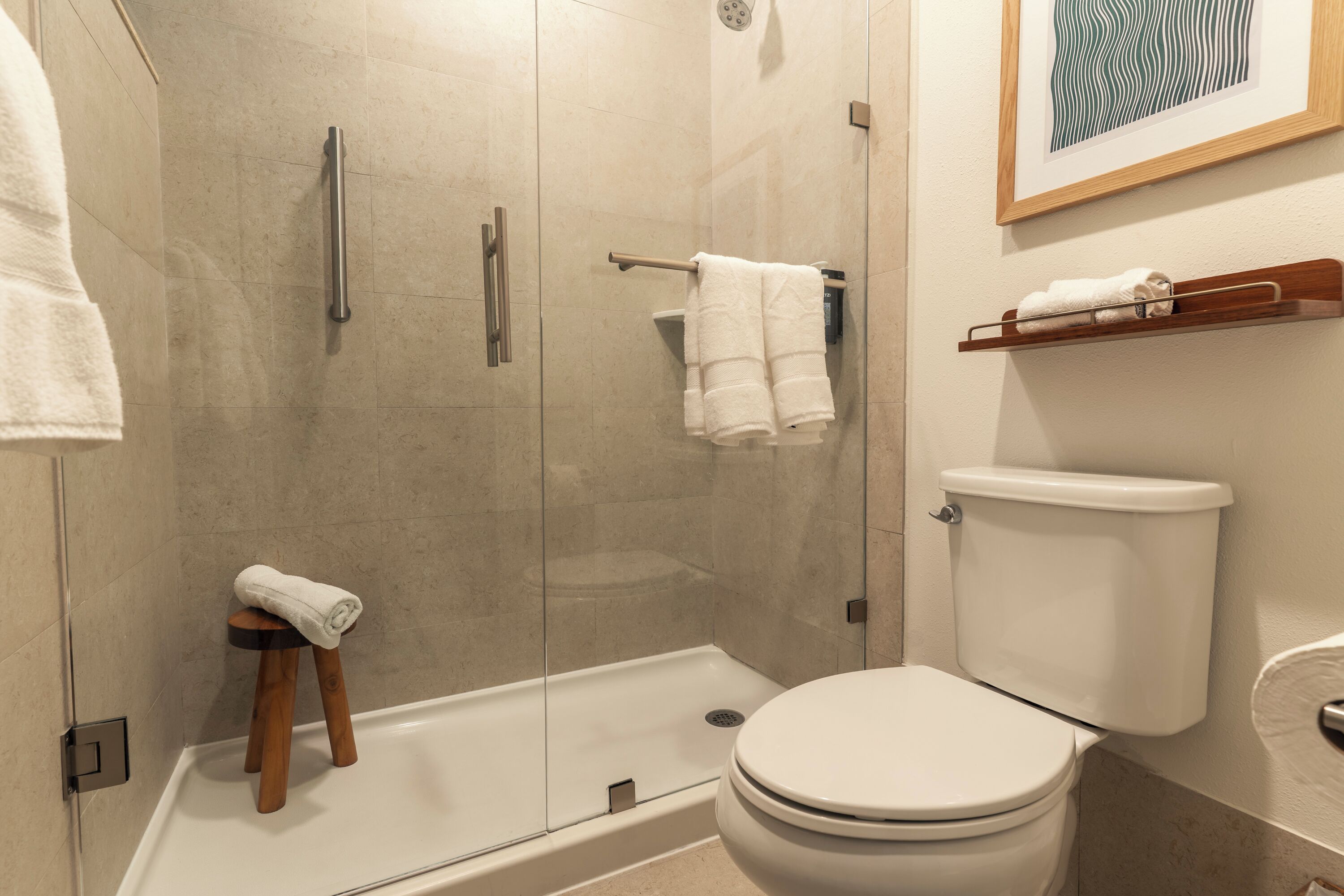 Un baño moderno con ducha acristalada, inodoro blanco y un taburete de madera con una toalla.