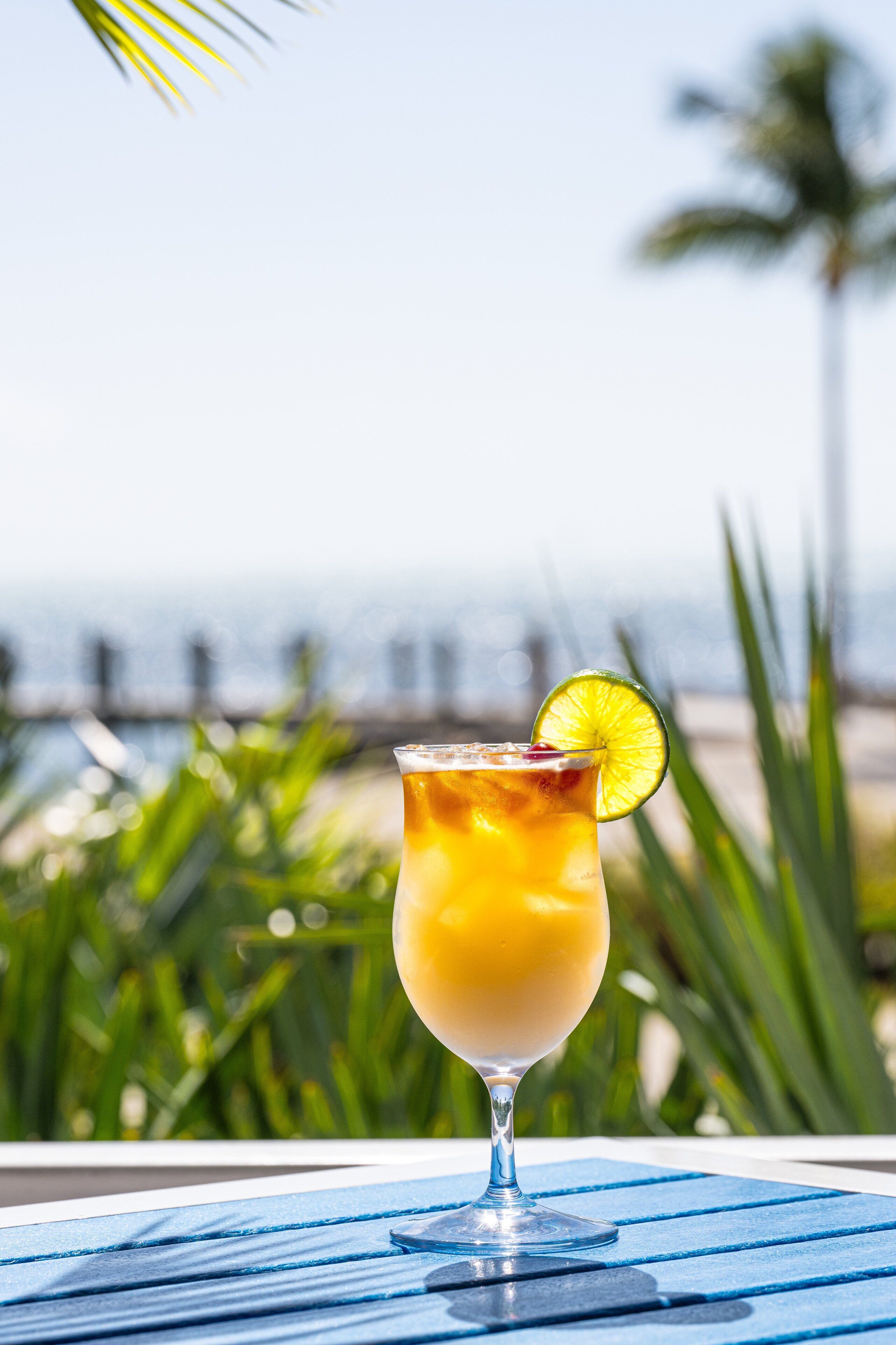 A cocktail glass with a lime garnish on a blue table, set against a beach backdrop with palm trees and ocean view.