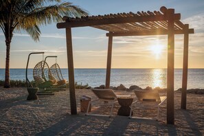 A beach setting with a wooden pergola, two wicker chairs, and a swing chair.