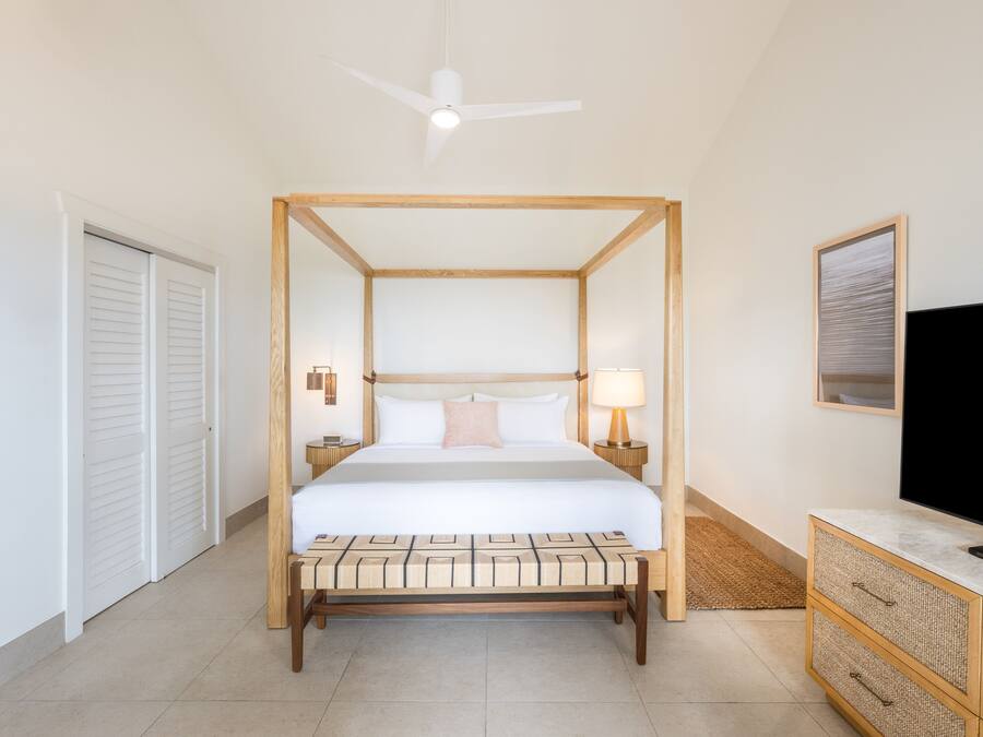 A four-poster bed with a wooden frame, a bedside table with a lamp, a television on a marble console, and a ceiling fan.
