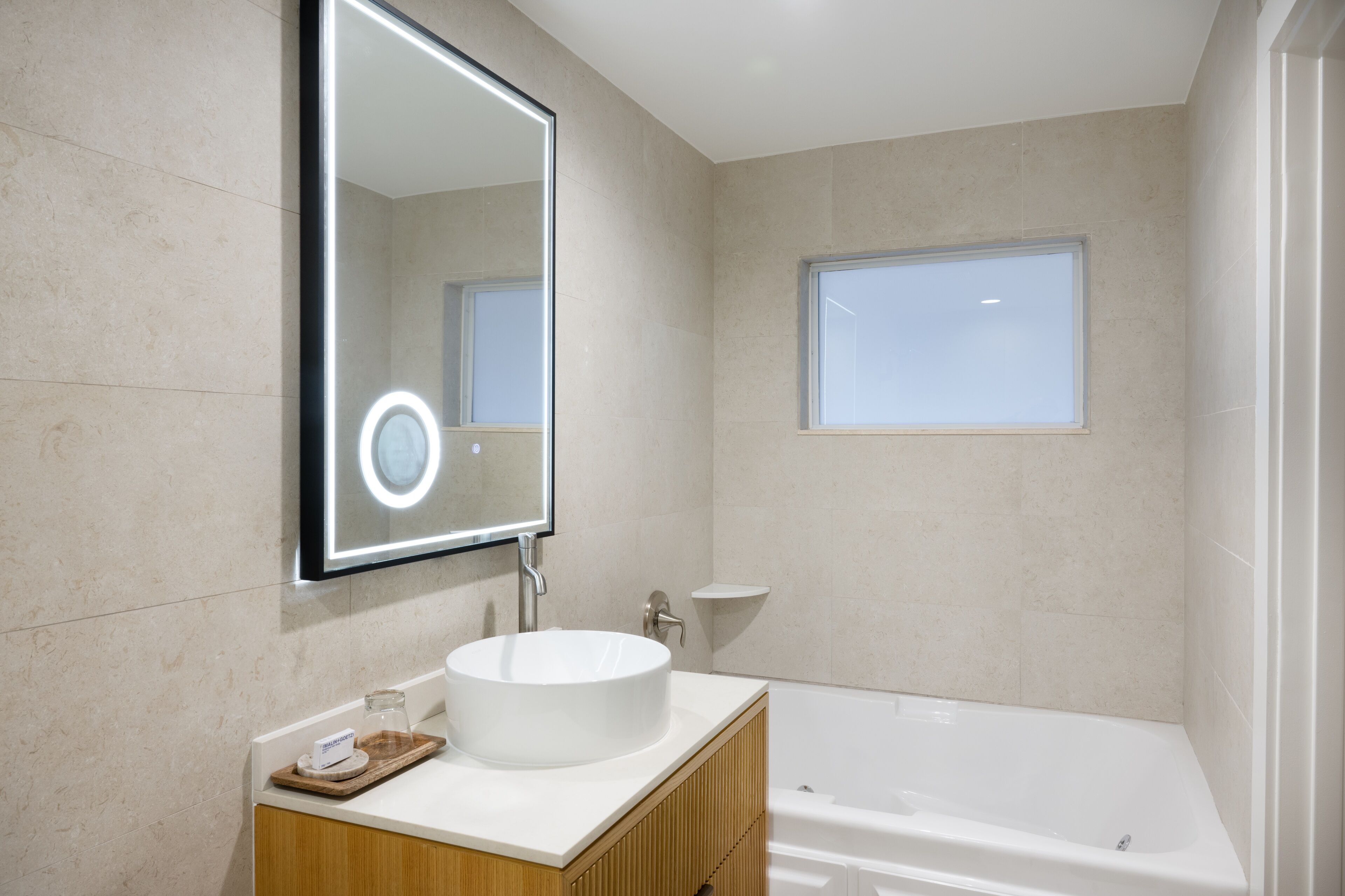A modern bathroom with a large mirror, a round sink, a wooden vanity, and a bathtub.