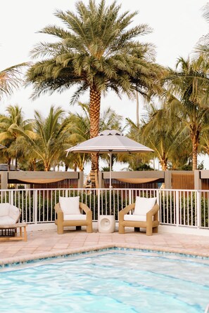 A pool area with two lounge chairs, a small umbrella, and palm trees.