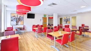 A modern dining area with red chairs and tables, a flat-screen TV mounted on the wall, and artwork on the walls. - Travelodge Aberdeen Central (Aberdeen)