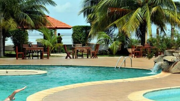 A pool with a person swimming, surrounded by palm trees and outdoor furniture. - Mariador Palace (Conakry)