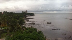 A coastal landscape with a beach, palm trees, and a distant building. - Mariador Palace (Conakry)