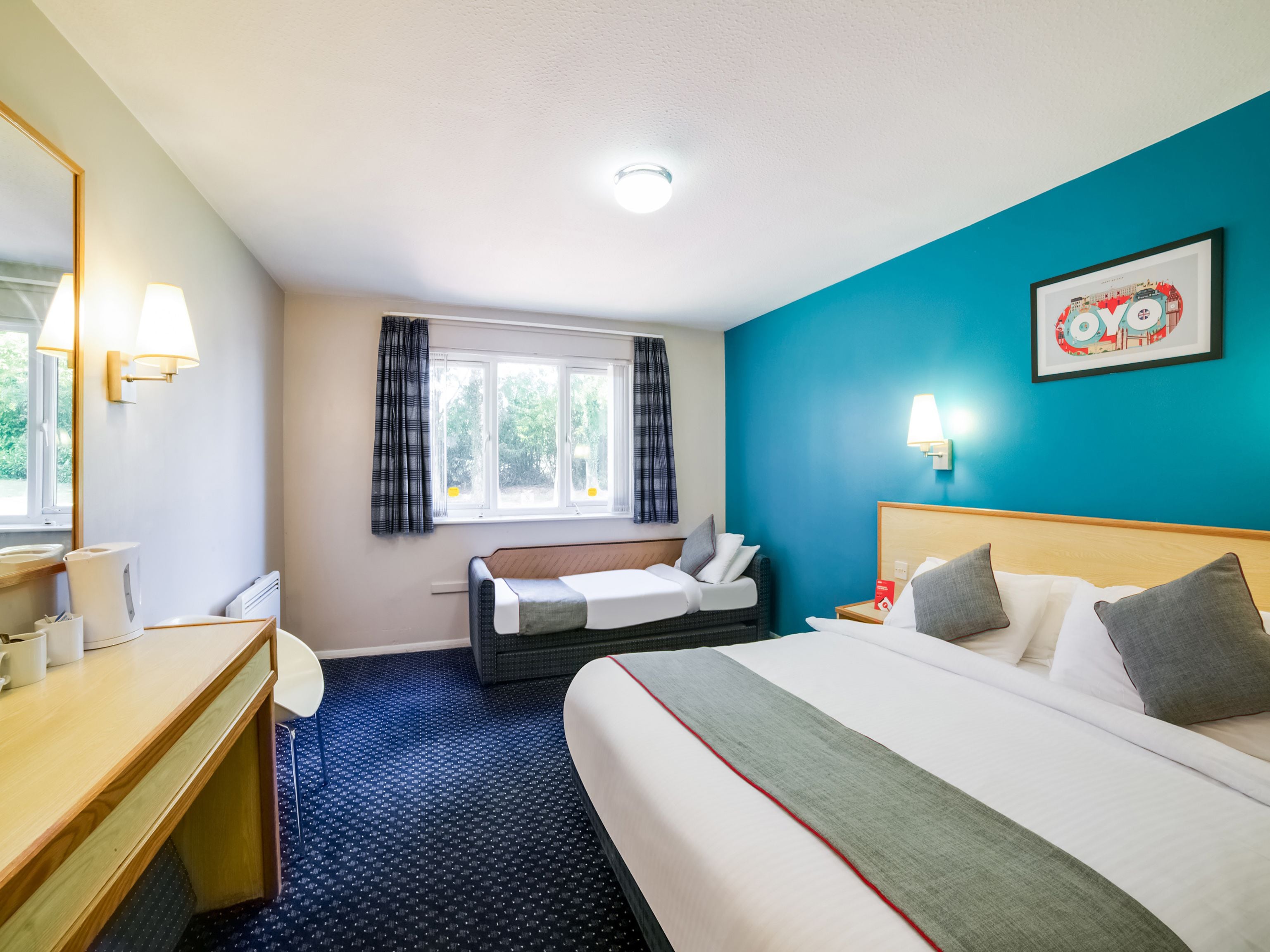 Une chambre d’hôtel comprenant un lit, un bureau, une chaise, une fenêtre avec des rideaux, une lampe fixée au mur et un tableau encadré.
