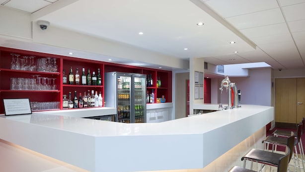 A modern bar area with a white countertop, red shelving with bottles and glasses, and a refrigerator. - Travelodge Glasgow Central (Glasgow)