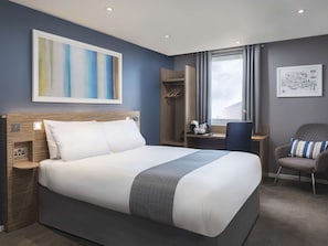 A modern hotel room with a large bed, a desk, a chair, and a window with a cityscape view.